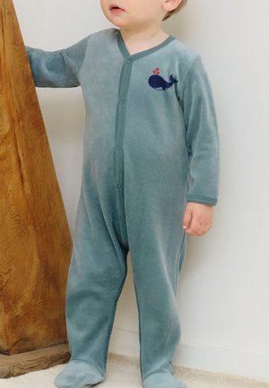 Toddler wearing blue onesie with whale design, standing and leaning on a wooden furniture piece against a white wall.
