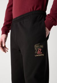 Zwarte joggingbroek van textuurstof met een elastische tailleband en zijzakken, voorzien van een gouden Lacoste-logo en krokodillenontwerp.