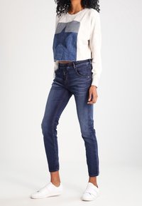 Sudadera corta de color gris claro con un diseño de estrella azul, combinada con jeans ajustados de mezclilla oscura y zapatillas blancas sin cordones.