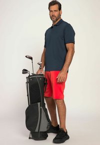 Herre golfbeklædning med en marineblå polo-shirt og røde shorts. Grå og sort golfbag med køller stående oprejst. Sorte sportsko.