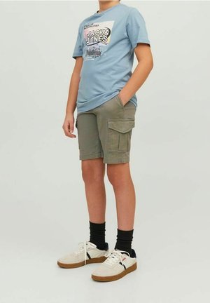 Dreng iført en blå Jack & Jones t-shirt, olivengrønne cargoshorts, sorte sokker og hvide sneakers, står med den ene hånd i lommen.