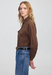 Brązowa koszula z długim rękawem, z kołnierzykiem i dwiema kieszeniami na piersi, zestawiona z jasnoniebieskimi dżinsami z wysokim stanem i czarnym paskiem.