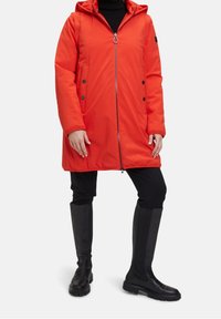Manteau rouge rembourré avec capuche, fermeture éclair sur toute la longueur, poches latérales et accents de boutons noirs. Porté sur un pantalon noir et des bottes noires jusqu'aux genoux.