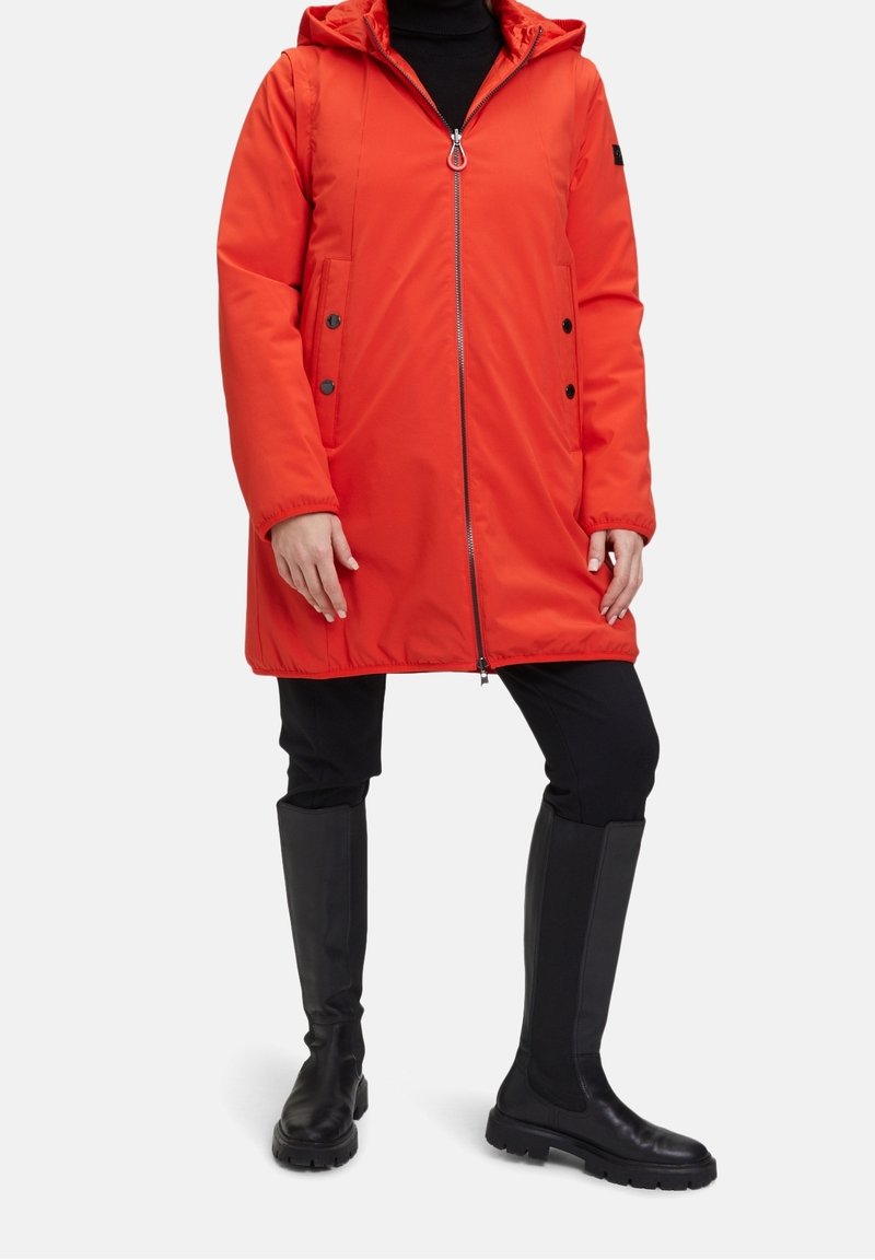 Manteau rouge rembourré avec capuche, fermeture éclair sur toute la longueur, poches latérales et accents de boutons noirs. Porté sur un pantalon noir et des bottes noires jusqu'aux genoux.