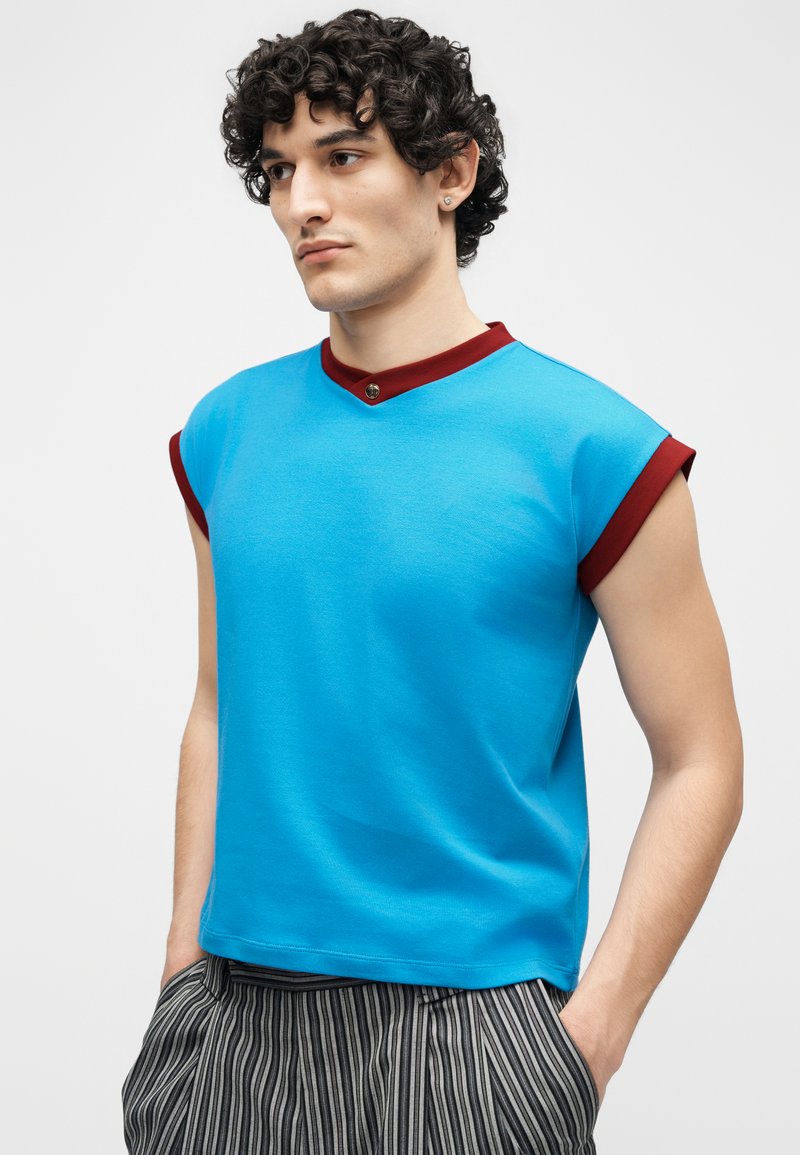 Young man with curly hair wearing a blue sleeveless top with maroon trim and striped pants, standing with hands in pockets.