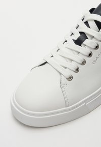 Adidas alb din piele cu vârf rotunjit, talpă din cauciuc alb texturat, accente negre și șireturi plate împletite prin ochiuri argintii.