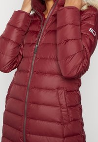 Abrigo acolchado color granate con cuello alto, cierre de cremallera y bolsillos laterales. Presenta una textura suave y detalles de pelo ligero en la capucha.