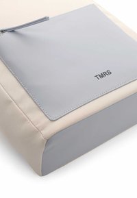 Bolso gris y beige con un acabado suave y mate, forma rectangular, bolsillo con cremallera y la marca "TMRS" en relieve en la parte frontal.