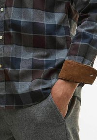 Geruit overhemd met knopen in marineblauw, grijs en bordeauxrood, met een bruine corduroy manchet. Close-upfoto met een grijze, gestructureerde broek.