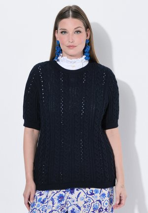 Kvinde med langt, glat hår iført navyblå kortærmet striktrøje, hvid bluse med højt halsudskæring, blå blomstret nederdel og store blå øreringe.