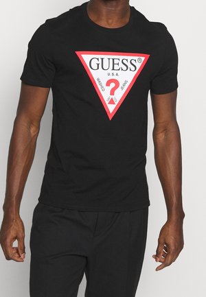 Mann trägt ein schwarzes T-Shirt mit einem großen roten und weißen dreieckigen Guess-Logo und schwarze Hosen vor einem einfarbigen hellen Hintergrund.