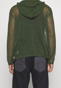 Felpa a rete verde con una vestibilità ampia, maniche lunghe e cappuccio. Notevole lavorazione a maglia texturizzata e orlo arrotondato. Abbinate a pantaloni scuri con dettagli curvi.