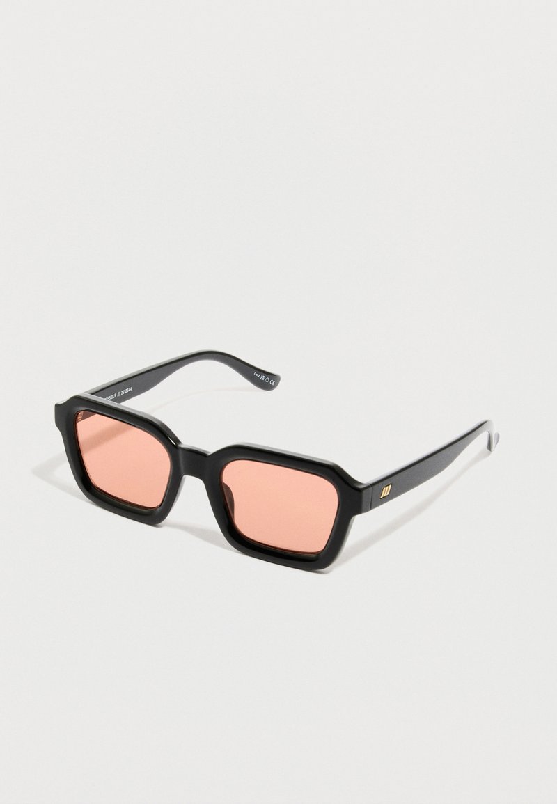 Μαύρα ορθογώνια γυαλιά ηλίου με παχιά πλαίσια και ροζ φακούς, τοποθετημένα πάνω σε λευκή επιφάνεια.