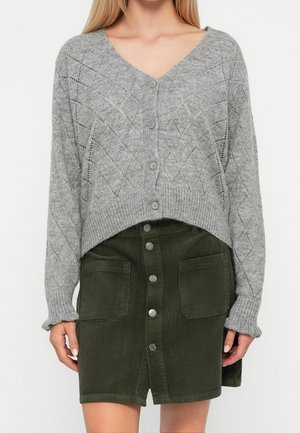 Woman wearing a gray knit buttoned cardigan with diamond patterns and a dark green corduroy buttoned skirt with front pockets.