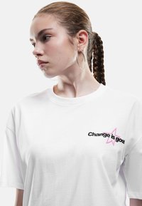 Ung kvinde med fletning, iført store hoop øreringe og en hvid T-shirt med teksten "Forandring er godt" og pink stjernedesign.