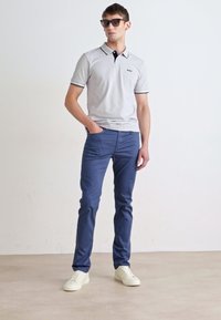 Polo shirt gris claro con ribete negro, combinado con pantalones azul marino y zapatillas blancas. Modelo usando gafas de sol, posando frente a un fondo liso.