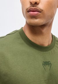 Κοντινό πλάνο ενός ατόμου που φοράει πουκάμισο ελιάς με σκούρο πράσινο λογότυπο φιδιού στο στήθος.