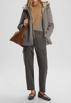 Cappotto grigio con cappuccio sopra un maglione beige, pantaloni grigi con tasche laterali e ballerine nere con borchie. Borsa a tracolla marrone appesa alla spalla.