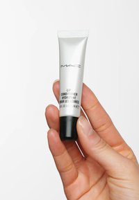 Silver tube av MAC Lip Conditioner, svart bas, med etiketttext i svart. Kapacitet på 15 ml, lätt klämd i en hand mot en vit bakgrund.