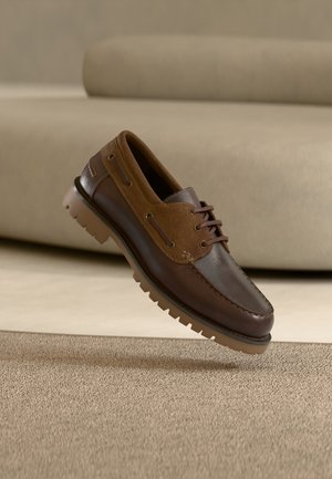 Καφέ δερμάτινο και σουέντ καθημερινό παπούτσι με κορδόνια και ανθεκτική σόλα, τοποθετημένο στον αέρα πάνω από μπεζ χαλι σε πατώματα και φόντο καναπέ.