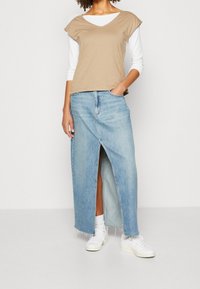 Top beige a maniche corte sovrapposto a una camicia bianca a maniche lunghe, abbinato a una gonna maxi in denim azzurro chiaro con uno spacco frontale e orlo sfilacciato. Sneakers bianche.
