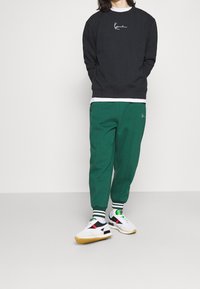 Sudadera negra con logo bordado en blanco, pantalones deportivos verdes y zapatillas blancas con detalles en negro y rojo y suelas de goma. Se ven calcetines a rayas.