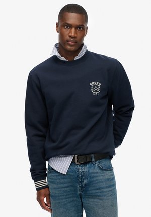 Sudadera azul marino con cuello redondo y logo bordado de "Super Dry". Presenta acentos a rayas en los puños, usada sobre una camisa con cuello.