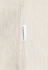 Gros plan d'un tissu en lin beige avec une couture verticale et une petite étiquette blanche cousue dans la couture affichant le nom de la marque Lindbergh.