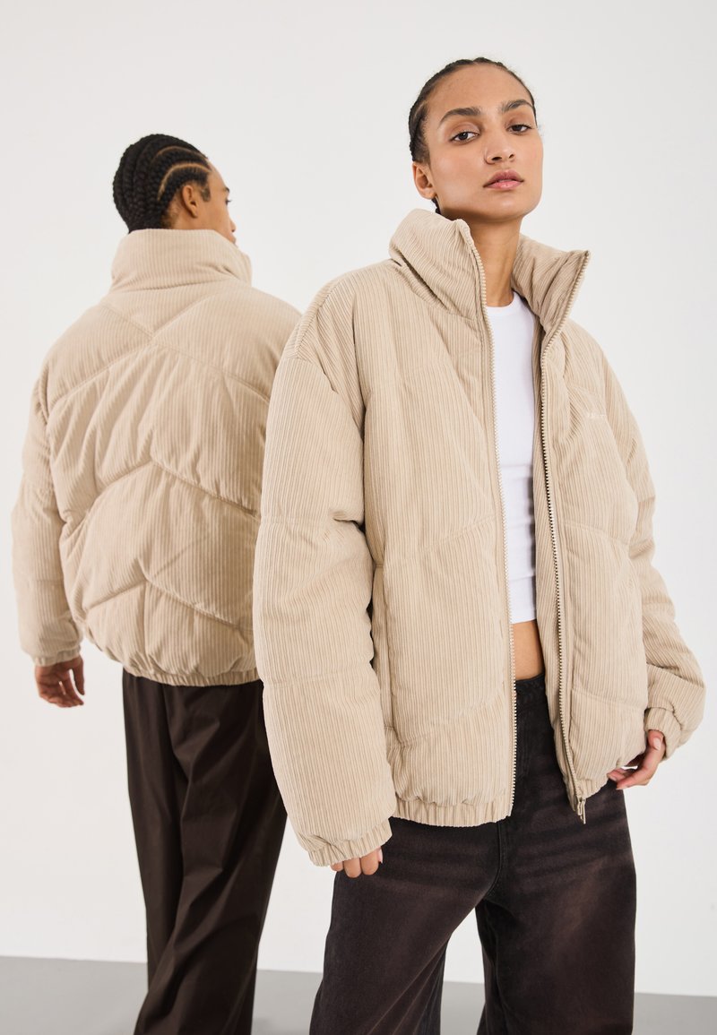 Beige kordfløyels-pufferjakke med høy krage, quiltet design og elastiske mansjetter, brukt med sorte bukser. Modell front- og bakfra.