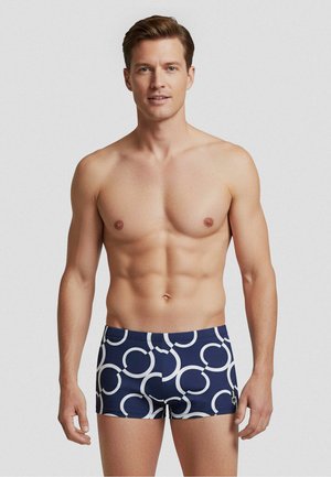 Fit man mit kurzen braunen Haaren trägt marineblaue Badehose mit großen weißen runden Mustern und steht vor einem schlichten Hintergrund.