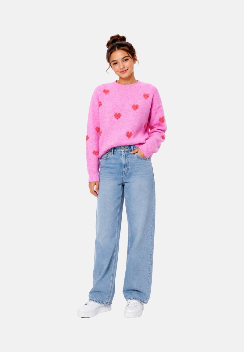 Jeune femme debout portant un pull rose avec des cœurs rouges, un jean large bleu clair et des baskets blanches, une main dans la poche.