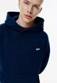 Felpa blu navy con tasca a canguro e logo ricamato bianco sul lato sinistro del petto. Realizzata in un tessuto morbido e strutturato con cappuccio regolabile.