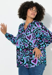 Frau mit lockigem Haar, die eine Brille trägt und ein buntes Langarmshirt mit blauen und violetten Herzmustern, posiert vor einer weißen Wand.