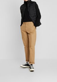 Czarna pikowana kamizelka na czarnym golfie, w połączeniu z beżowymi spodniami chino i czarnymi trampkami. Wąski krój o swobodnym wyglądzie.