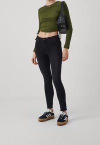 Jeans negros ajustados de cintura alta, combinados con una blusa corta de manga larga verde y zapatillas negras con detalles blancos y suelas de goma.