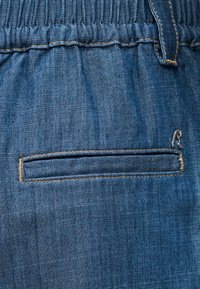 Prim-plan al unui material din denim albastru cu o talie elastică și un buzunar mic orizontal cu cusături contrastante.