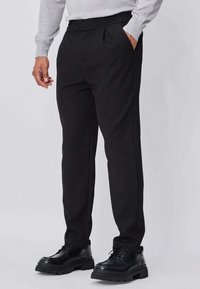 Pantalon noir en tissu lisse, avec une coupe ajustée, une ceinture élastique et des poches latérales, assorti à des chaussures noires à lacets.