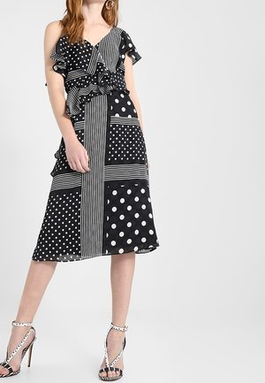 Kobieta ubrana w czarno-białą sukienkę z mieszanką kropek i pasków z falbanami, do której dobrano pasujące sandały na wysokim obcasie.