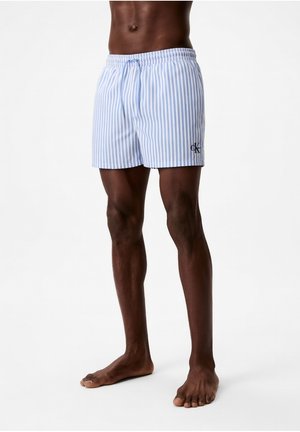 Modelo masculino con bañador corto de rayas verticales azul claro y blanco con logo CK en el muslo izquierdo, de pie descalzo sobre fondo blanco.