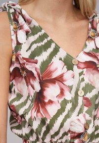 Blouse florale à col en V, ornée de fleurs roses et bourgognes sur un fond à rayures vertes. Comprend des boutons en bois et un détail de lien aux épaules.