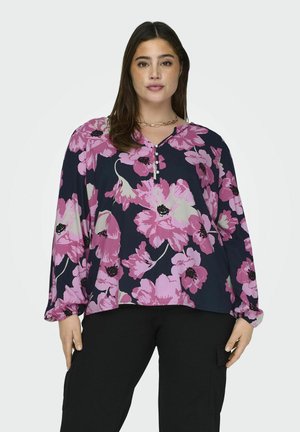 Blouse florale avec un fond bleu marine et de grandes fleurs roses, un décolleté en V et un détail de bouton au niveau de la poitrine. Manches longues, coupe ample.