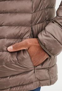 Braune Pufferjacke mit diagonaler Steppung, Reißverschluss-Seitentasche und weichem Material, das mit einer kontrastierenden Innenauskleidung sichtbar ist.