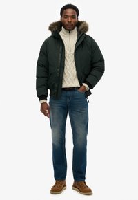 Zielona kurtka puchowa z kapturem wyłożonym futrem, noszona na kremowym swetrze w warkoczowy splot. Do tego niebieskie dżinsy i brązowe buty.