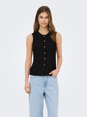 Woman wearing a sleeveless black buttoned top and light blue high-waisted jeans, standing with one hand in pocket against white background.