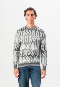 Maglione grigio a maglia con grande logo ripetuto "GUESS" in nero, collo giro e polsini e orlo a costine. Indossato con jeans blu.
