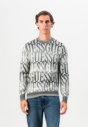 Szara dzianinowa bluza z dużym, powtarzającym się logo "GUESS" w kolorze czarnym, z okrągłym dekoltem i ściągaczami przy mankietach oraz u dołu. Noszona z niebieskimi dżinsami.