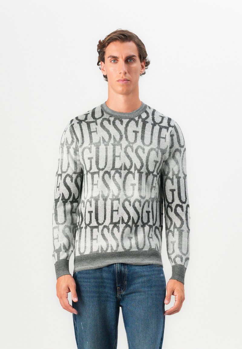 Maglione grigio a maglia con grande logo ripetuto "GUESS" in nero, collo giro e polsini e orlo a costine. Indossato con jeans blu.