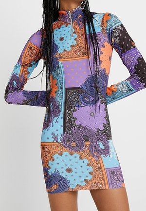 Femme portant une robe mini colorée à manches longues avec un col montant et une fermeture éclair, présentant un motif patchwork de dragons et de motifs paisley.