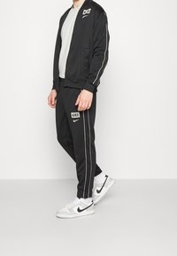 Tuta Nike nera con dettagli bianchi, giacca con zip e toppa con logo, e pantaloni aderenti abbinati. Indossata con sneakers bianche e grigie.