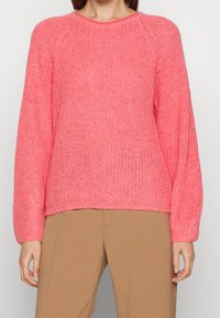 Frau in einem rosa gestrickten Langarmpullover und hellbraunen Hosen, die mit entspannten Armen an ihrer Seite steht.